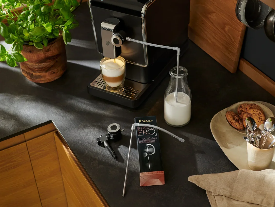 Ein »Esperto Pro« Tchibo Kaffeevollautomat bereitet Kaffee in einem Glas zu. Daneben stehen eine Milchflasche und Zubehör.