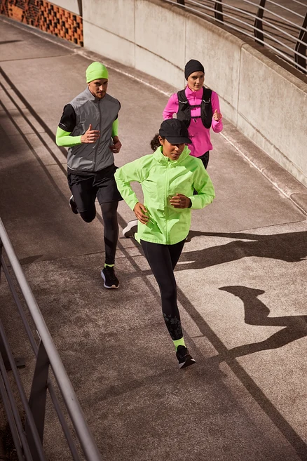 Drei Läufer, zwei Frauen und ein Mann, laufen auf einem Betonweg. Die Frau vorne trägt eine schwarze Sport-Cap, eine neonlime Laufjacke, schwarze Sportleggings »ActivePower« und neonfarbene Laufsocken. Der Mann in der Mitte trägt eine neonlime Thermomütze, eine reflektierende Laufweste über einem schwarzen Sportshirt und neonlime Lauf-Armlinge, schwarze Sportshorts über Thermo-Sporttights und neonfarbene Laufsocken. Die Frau hinten trägt eine schwarze Thermomütze, ein rosa Thermo-Sportshirt mit Half-Zip und eine Laufweste.
