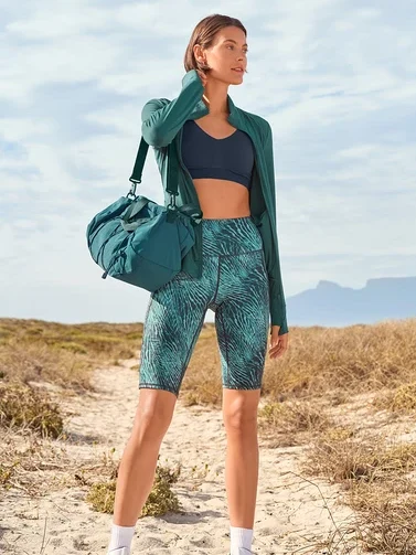 Frau mit Sportbekleidung und grüner Sporttasche auf dem Weg. Sie trägt Shorts mit Blattmuster, Sport-BH und eine Jacke.