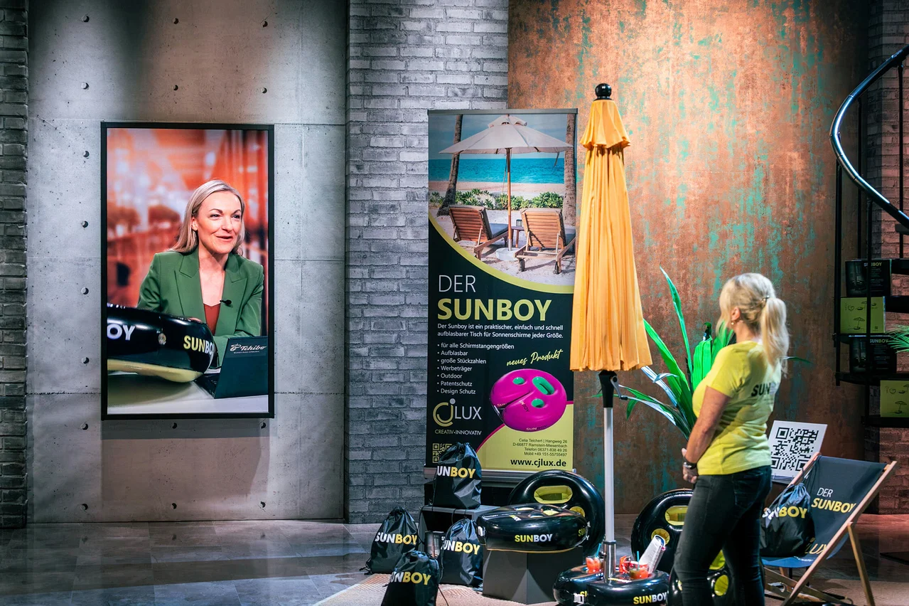 Frau in gelbem Shirt steht neben gelbem Sonnenschirm. Im Hintergrund ist ein Bildschirm mit einer Frau zu sehen. Mehrere Sunboy Produkte sind zu sehen.