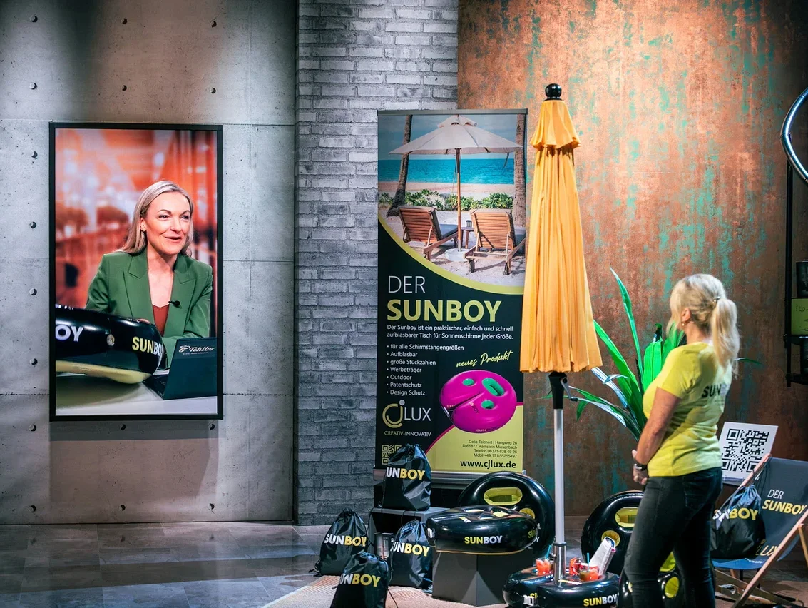 Frau in gelbem Shirt steht neben gelbem Sonnenschirm. Im Hintergrund ist ein Bildschirm mit einer Frau zu sehen. Mehrere Sunboy Produkte sind zu sehen.