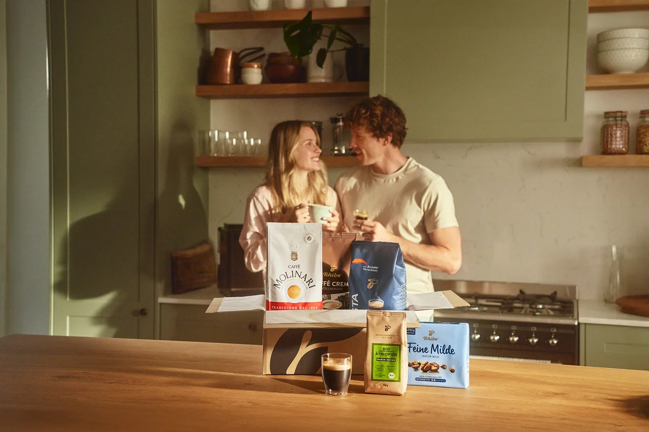 Zwei Personen stehen in einer Küche und halten Kaffeetassen. Vor ihnen steht eine offene Kiste mit verschiedenen Kaffeesorten, darunter Molinari und Tchibo.