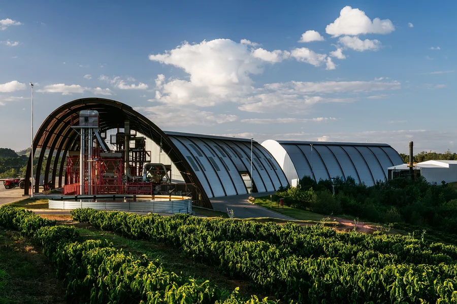 Reihen grüner Kaffeepflanzen im Vordergrund führen zu einer modernen Industrieanlage mit gewölbten, hangarähnlichen Gebäuden im Hintergrund. Rote Verarbeitungsmaschinen sind im linken Gebäude unter blauem Himmel mit weißen Wolken sichtbar.