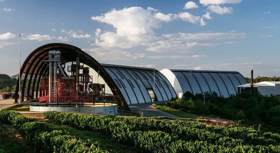 Reihen grüner Kaffeepflanzen im Vordergrund führen zu einer modernen Industrieanlage mit gewölbten, hangarähnlichen Gebäuden im Hintergrund. Rote Verarbeitungsmaschinen sind im linken Gebäude unter blauem Himmel mit weißen Wolken sichtbar.