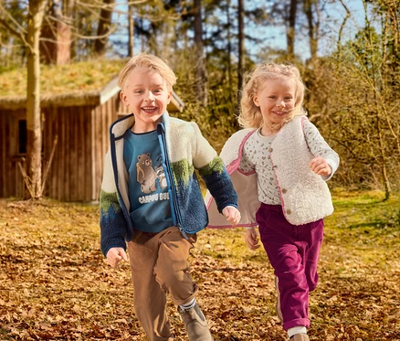 Zwei lächelnde Kinder, ein Junge und ein Mädchen, laufen durch eine laubbedeckte Naturlandschaft. Der Junge trägt ein blaues Shirt mit Motiv, eine farbige Fleecejacke und braune Cordhosen. Das Mädchen trägt ein gemustertes Shirt, eine cremefarbene flauschige Weste und magentafarbene Cordhosen. Im Hintergrund ist ein Holzhaus mit Grasdach zu sehen.