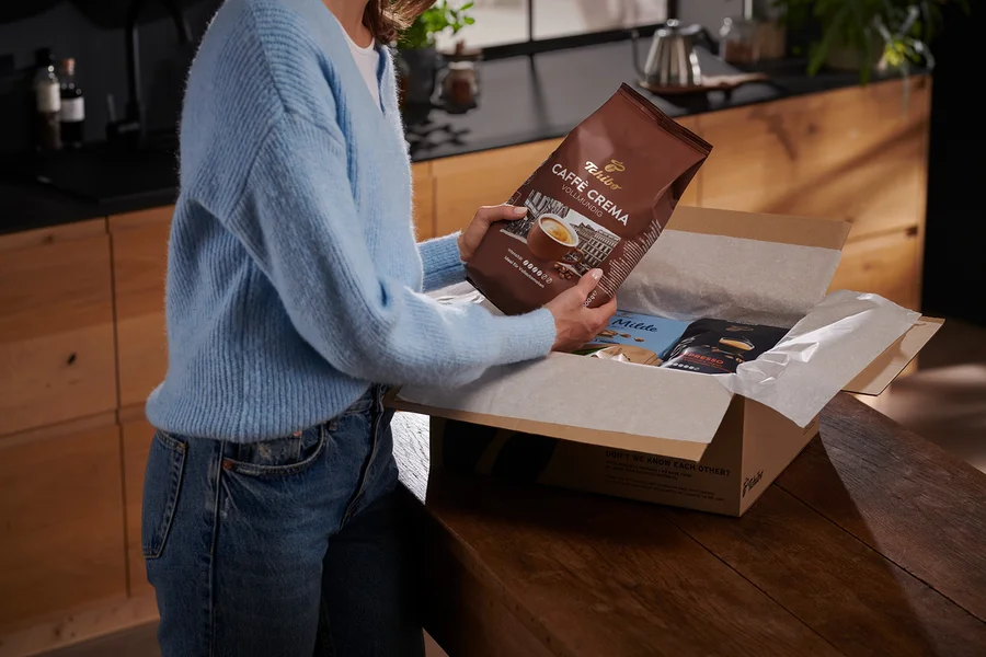 Frau hält eine Packung Tchibo Caffe Crema Kaffee über eine Schachtel mit anderen Kaffeepackungen.