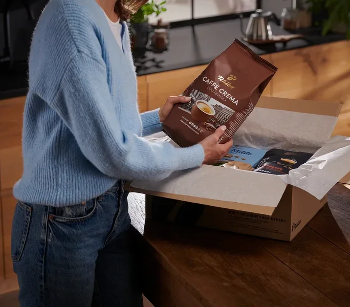 Frau hält eine Packung Tchibo Caffe Crema Kaffee über eine Schachtel mit anderen Kaffeepackungen.