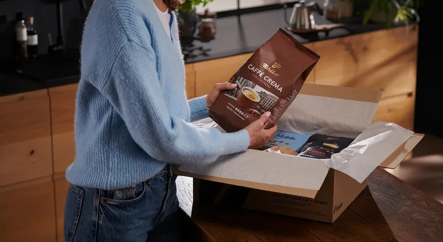 Frau hält eine Packung Tchibo Caffe Crema Kaffee über eine Schachtel mit anderen Kaffeepackungen.