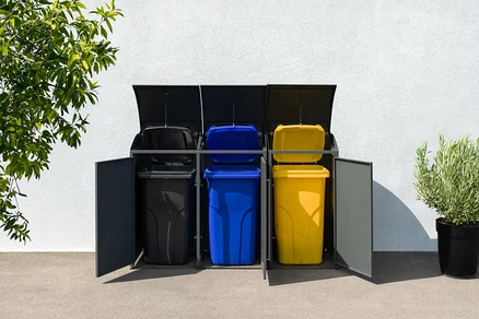 Drei farbige Mülltonnen in einer grauen Mülltonnenbox, 3er, stehen vor einer weißen Wand.
