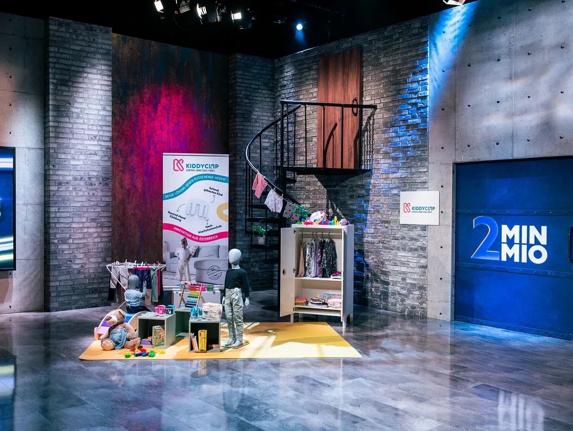 Auf einem Set mit Backsteinwänden stehen eine Wendeltreppe, ein Kleiderständer, Spielzeug und zwei Schaufensterpuppen.