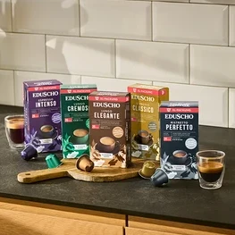 Fünf Packungen Eduscho Kaffeekapseln (Espresso Intenso, Lungo Cremoso, Lungo Elegante, Espresso Classico, Ristretto Perfetto) stehen auf einer Küchentheke mit zwei Tassen Kaffee und losen Kapseln.