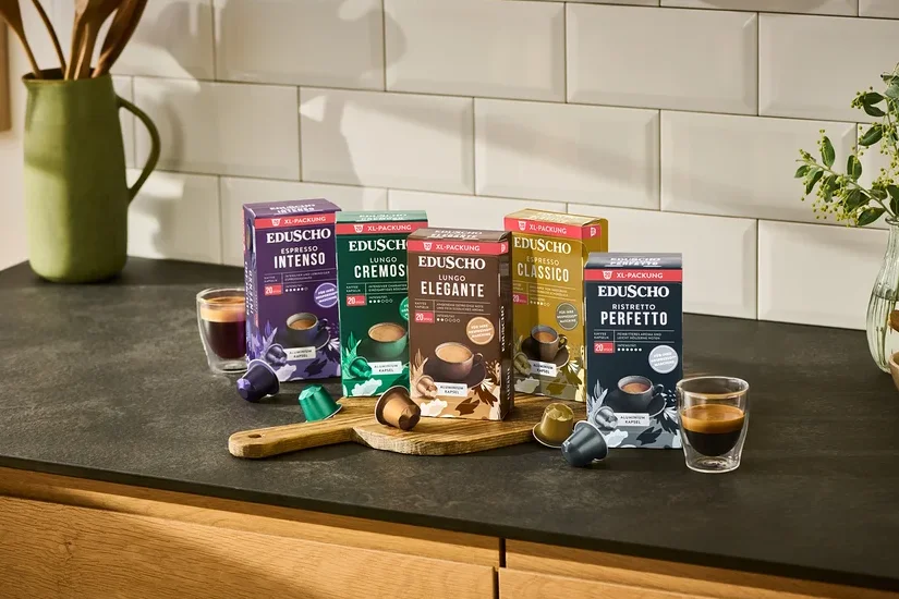 Fünf Packungen Eduscho Kaffeekapseln (Espresso Intenso, Lungo Cremoso, Lungo Elegante, Espresso Classico, Ristretto Perfetto) stehen auf einer Küchentheke mit zwei Tassen Kaffee und losen Kapseln.