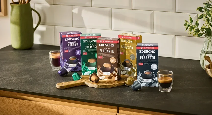 Fünf Packungen Eduscho Kaffeekapseln (Espresso Intenso, Lungo Cremoso, Lungo Elegante, Espresso Classico, Ristretto Perfetto) stehen auf einer Küchentheke mit zwei Tassen Kaffee und losen Kapseln.