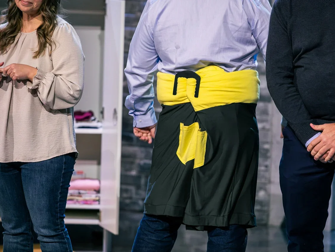 Drei Personen stehen vor einem offenen Schrank. Die mittlere Person trägt eine gelbe Schärpe und eine schwarze Schürze.