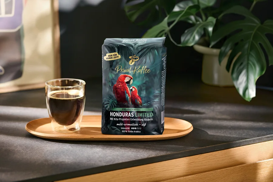 Ein dunkelgrüner Kaffeebeutel „Privat Kaffee Honduras Limited“ mit zwei roten Aras und eine Glastasse mit schwarzem Kaffee stehen auf einem Holztablett. Der Beutel trägt Aufschriften wie „NUR FÜR KURZE ZEIT!“ und „100% Tchibo Arabica“. Im Hintergrund ist eine grüne Pflanze zu sehen.