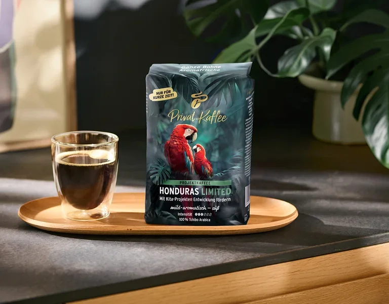 Ein dunkelgrüner Kaffeebeutel „Privat Kaffee Honduras Limited“ mit zwei roten Aras und eine Glastasse mit schwarzem Kaffee stehen auf einem Holztablett. Der Beutel trägt Aufschriften wie „NUR FÜR KURZE ZEIT!“ und „100% Tchibo Arabica“. Im Hintergrund ist eine grüne Pflanze zu sehen.