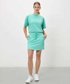 Frau steht mit Händen in den Taschen eines türkisfarbenen Rocks, dazu passendes T-Shirt und weiße Turnschuhe.