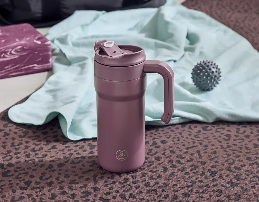 Eine lila Thermoskanne mit Griff steht auf einer braunen Yogamatte mit Leopardenmuster. Daneben liegen ein grauer Igelball, ein türkisfarbenes Handtuch und ein lila Yogablock.