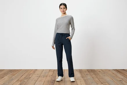 Frau mit grau-weiß gestreiftem Langarmshirt und marineblauer Hose mit Händen in den Taschen, vor weißem Hintergrund.
