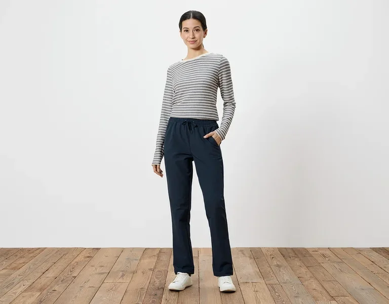 Frau mit grau-weiß gestreiftem Langarmshirt und marineblauer Hose mit Händen in den Taschen, vor weißem Hintergrund.