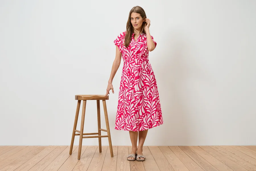 Frau im pinkfarbenen Kleid mit weißem Blattmuster, stehend neben einem Holzschemel.