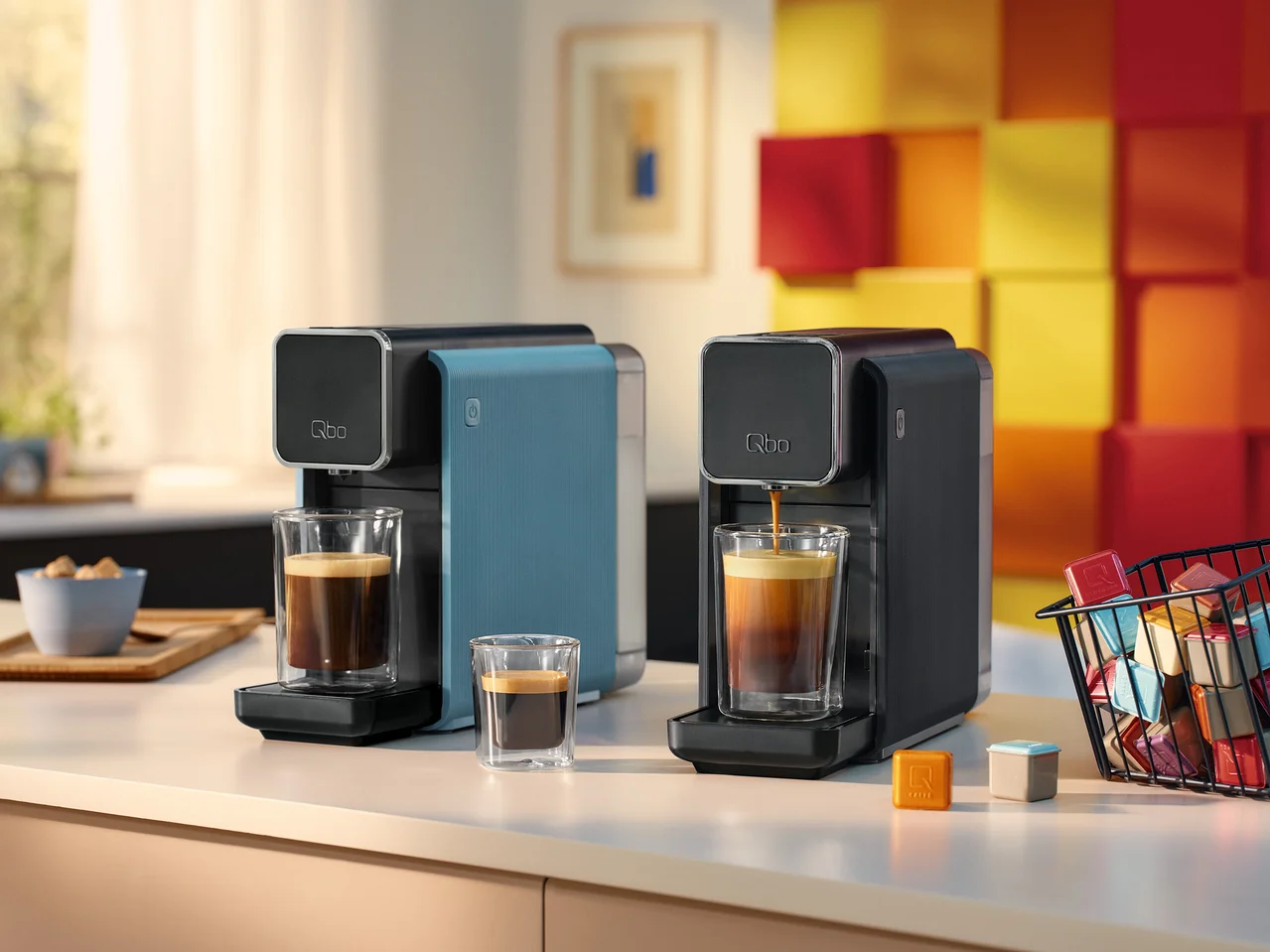 Zwei Qbo-Kaffeemaschinen, eine blaue und eine schwarze, mit Kaffee in Gläsern. Kapseln in einem Korb.