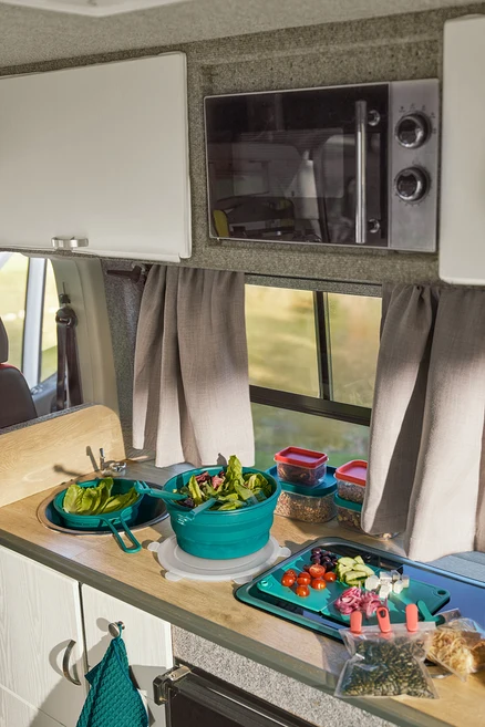 Küchenzeile im Camper mit Platzspar-Sieb im Spülbecken, Faltbarer Salatschüssel mit Salat und 2-in-1-Pfannenwender-und-Zange. Auf dem Faltbaren Schneidebrett liegen geschnittene Zutaten. Daneben sind zwei Aromaclips und zwei Aufbewahrungsdosen. Eine Mikrowelle hängt über dem Fenster.