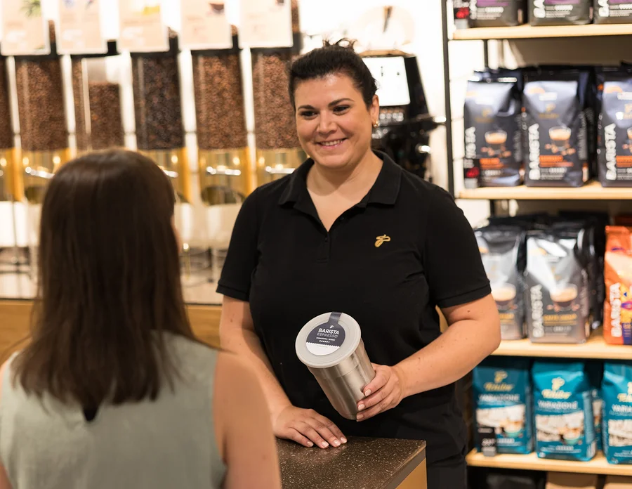 Lächelnde Frau in schwarzem Poloshirt hält einen silbernen Behälter mit „BARISTA ESPRESSO“-Etikett und interagiert mit einer Kundin an einer Theke mit Kaffeeprodukten im Hintergrund.