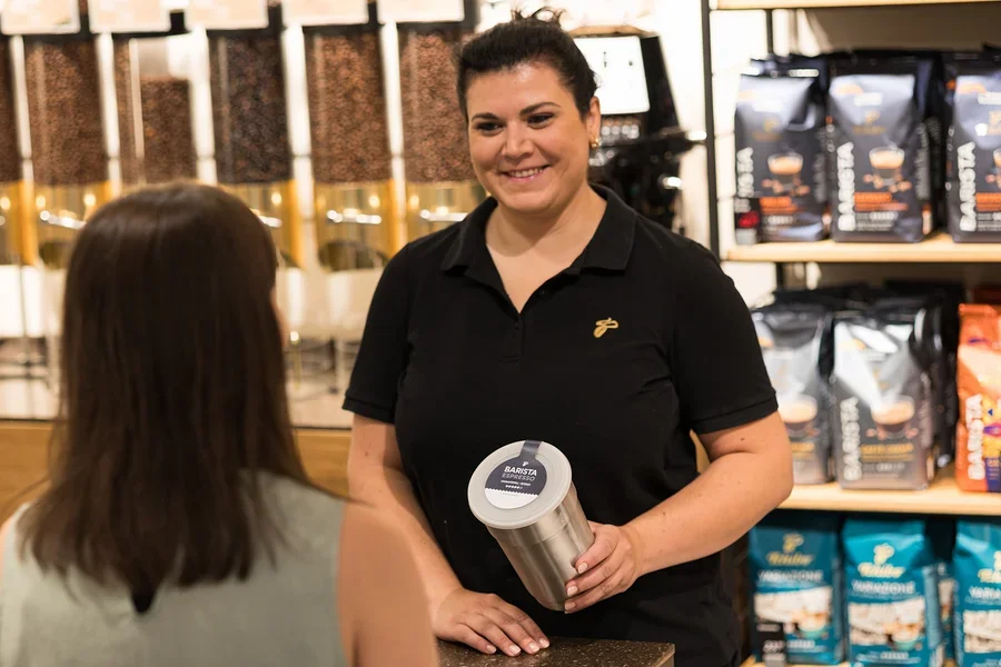 Lächelnde Frau in schwarzem Poloshirt hält einen silbernen Behälter mit „BARISTA ESPRESSO“-Etikett und interagiert mit einer Kundin an einer Theke mit Kaffeeprodukten im Hintergrund.