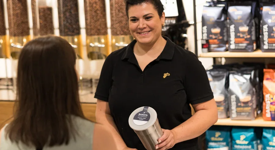 Lächelnde Frau in schwarzem Poloshirt hält einen silbernen Behälter mit „BARISTA ESPRESSO“-Etikett und interagiert mit einer Kundin an einer Theke mit Kaffeeprodukten im Hintergrund.