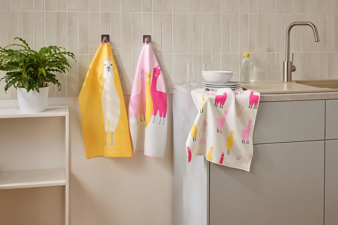 Drei Geschirrtücher mit Lama-Motiven hängen in einer Küche neben einem Waschbecken und einem Regal.