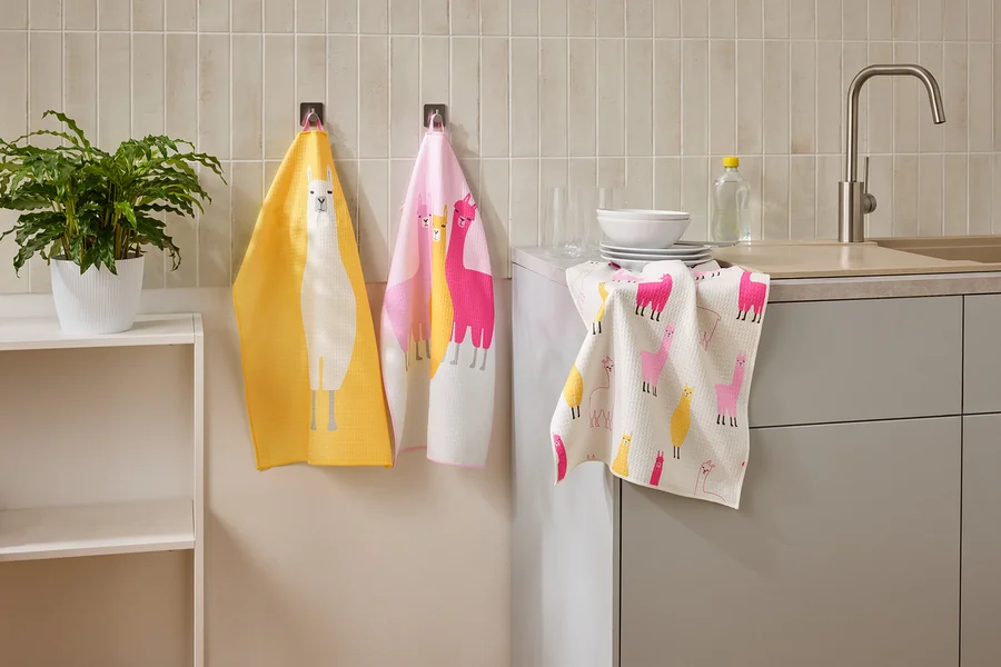 Drei Geschirrtücher mit Lama-Motiven hängen in einer Küche neben einem Waschbecken und einem Regal.