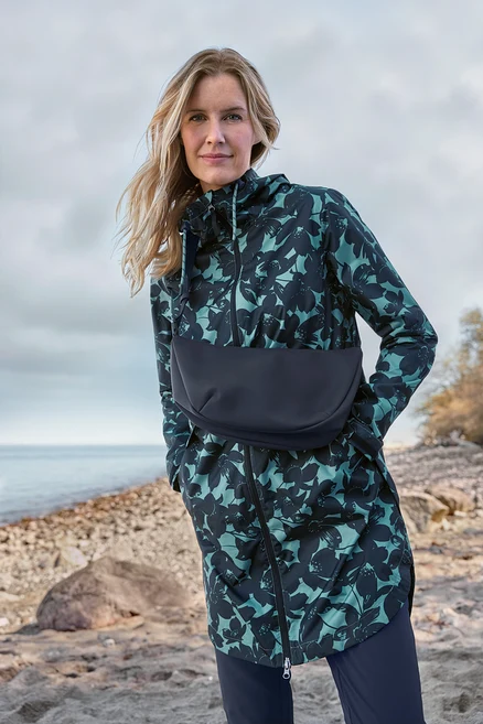 Frau mit blondem Haar trägt eine dunkelpetrolfarbene und schwarze Kapuzenjacke mit Blumenmuster und schwarzem Einsatz, stehend an einem felsigen Strand am Meer.