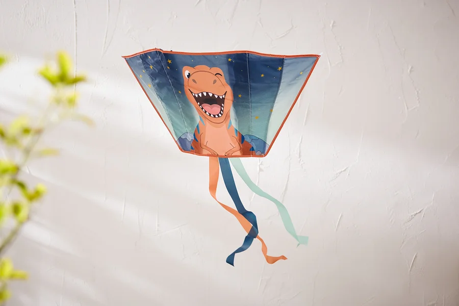 Ein trapezförmiger Drachen mit einem lachenden orangefarbenen Dinosaurier und Sternen hängt an einer strukturierten weißen Wand. Unten hängen orange, dunkelblaue und hellblaue Bänder.