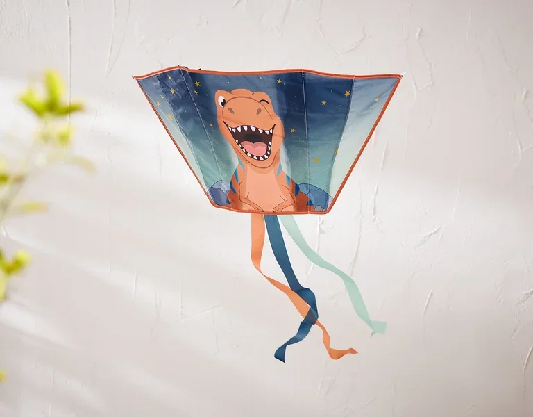 Ein trapezförmiger Drachen mit einem lachenden orangefarbenen Dinosaurier und Sternen hängt an einer strukturierten weißen Wand. Unten hängen orange, dunkelblaue und hellblaue Bänder.