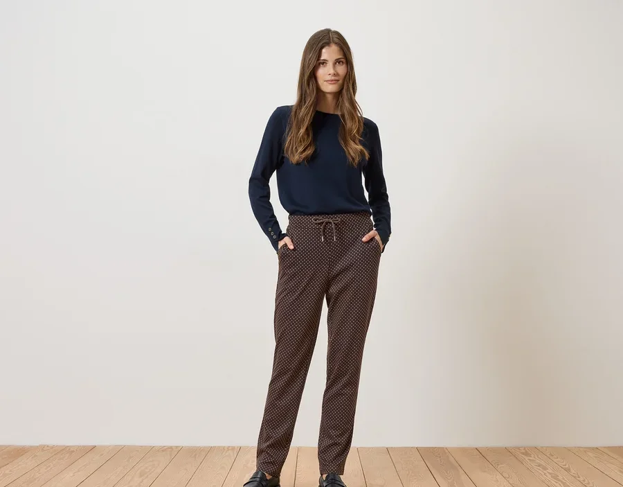 Eine Frau steht vor einer weißen Wand und trägt einen marineblauen Pullover, gemusterte Hose und schwarze Schuhe.