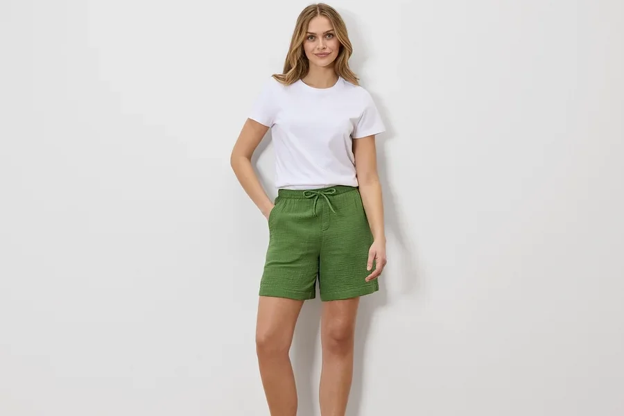 Frau in weißem T-Shirt und grünen Shorts mit Kordelzug, stehend vor einer weißen Wand.