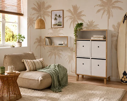 Ein entspanntes Zimmer mit Palmen-Tapete und einem zotteligen Teppich, mit einem beigen Cord-Sessel mit gestreiftem Kissen und grüner Decke, einem Rattan-Beistelltisch und einem Holzregal mit weißen Aufbewahrungsboxen. Ein gerahmtes Bild von Palmen und einem Vintage-Van hängt über einem schwebenden Regal, und ein Surfbrett lehnt an der Wand. Ein Fenster mit einem Bambusrollo zeigt Grün im Freien.