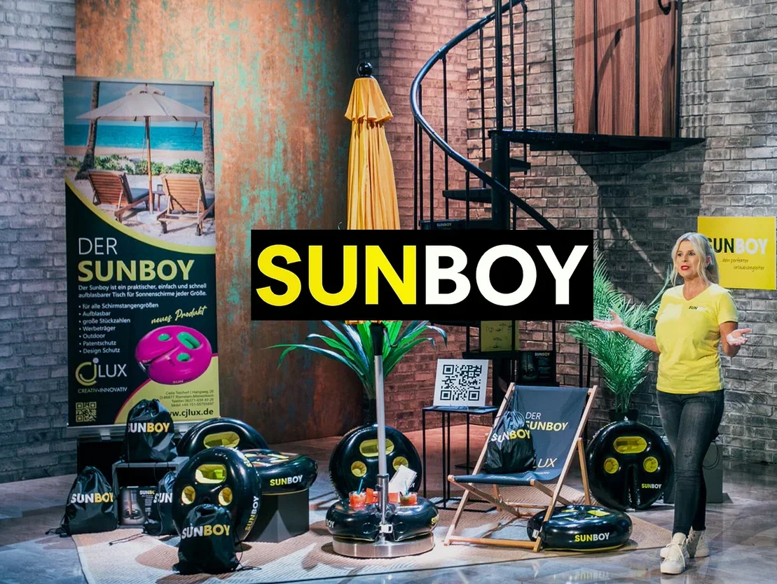 Frau präsentiert Sunboy-Produkte: aufblasbare Tische, Sonnenschirmständer, Strandstuhl und Tragetaschen vor Backsteinmauer und Wendeltreppe.