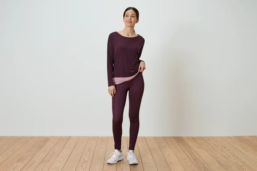 Frau steht und trägt ein langärmliges, auberginefarbenes Hemd und auberginefarbene Leggings.