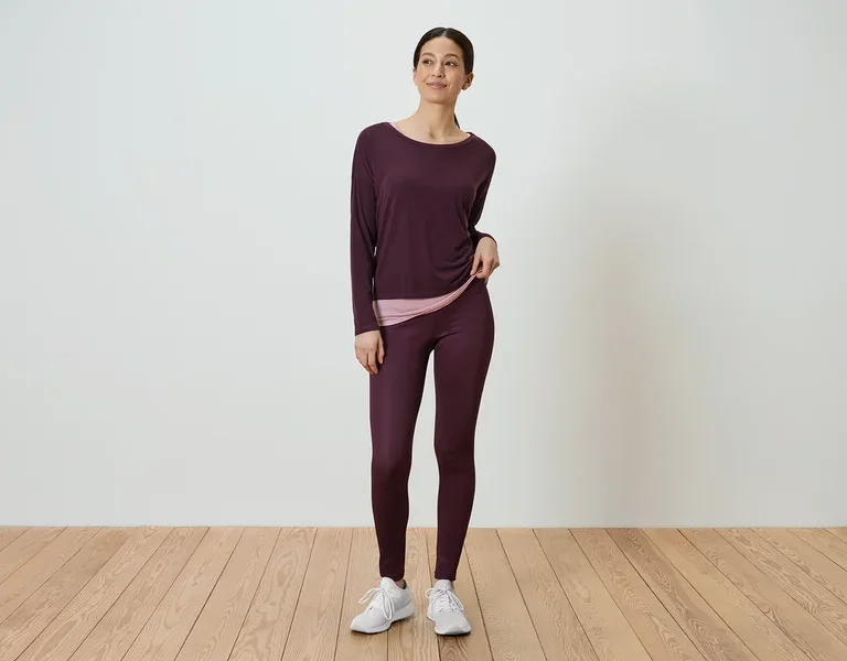 Frau steht und trägt ein langärmliges, auberginefarbenes Hemd und auberginefarbene Leggings.