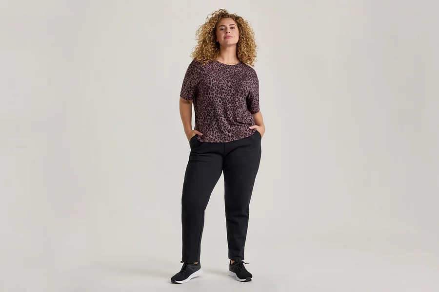 Eine Frau mit lockigem Haar steht frontal, trägt ein kurzärmeliges, dunkelviolettes Leopardenmuster-Oberteil, schwarze Hosen und schwarze Turnschuhe. Ihre Hände stecken in den Hosentaschen.