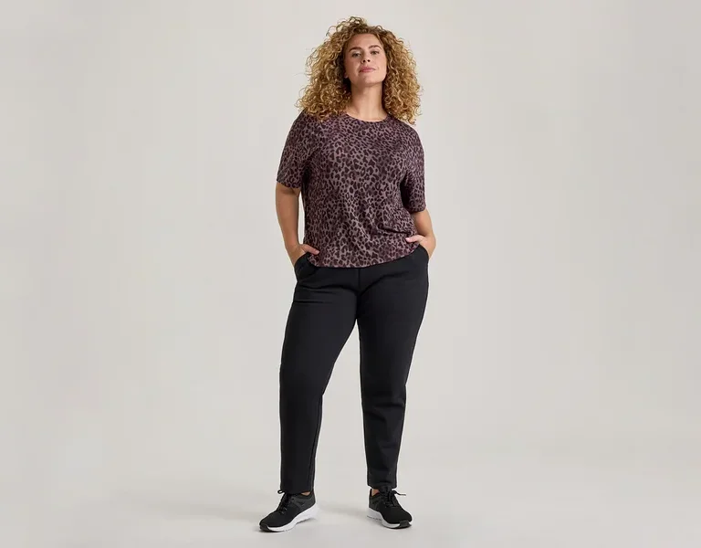 Eine Frau mit lockigem Haar steht frontal, trägt ein kurzärmeliges, dunkelviolettes Leopardenmuster-Oberteil, schwarze Hosen und schwarze Turnschuhe. Ihre Hände stecken in den Hosentaschen.