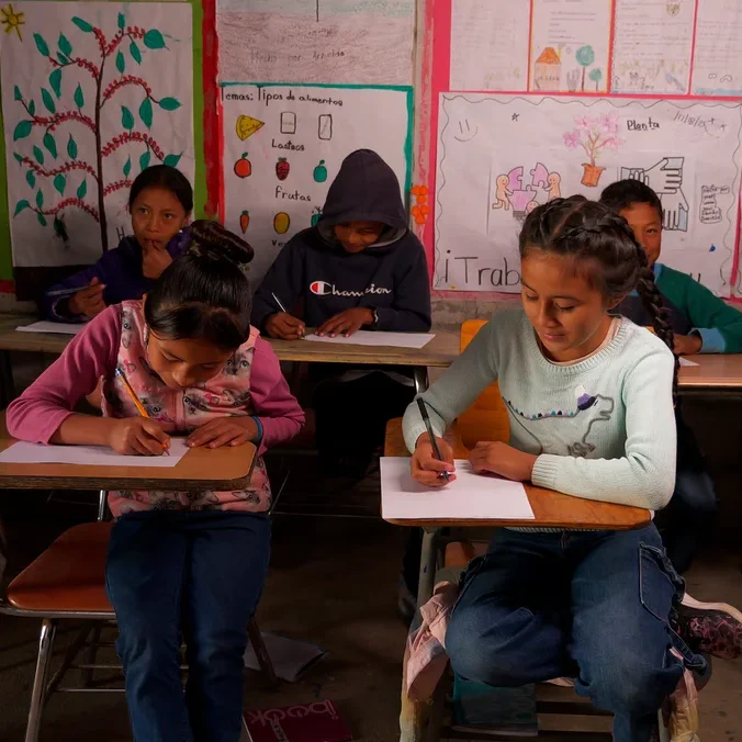 Mehrere Kinder sitzen in einem Klassenzimmer an Holztischen und schreiben auf Papier. Zwei Mädchen im Vordergrund konzentrieren sich auf das Schreiben, eines in einer rosa Weste und das andere in einem hellblauen Pullover. Die Wände des Klassenzimmers sind mit Lernplakaten dekoriert.