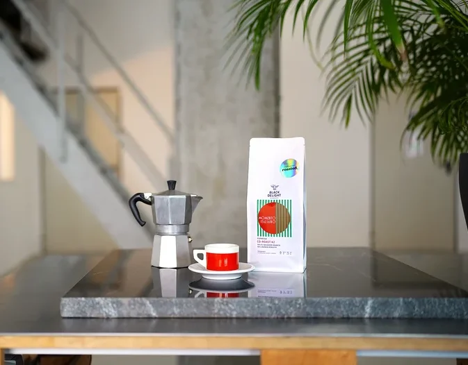 Auf einem Tisch stehen eine Kaffeemaschine, eine Tasse und eine Packung Black Delight x roasted. - Momento Italiano Co-Roast #2 Espresso.