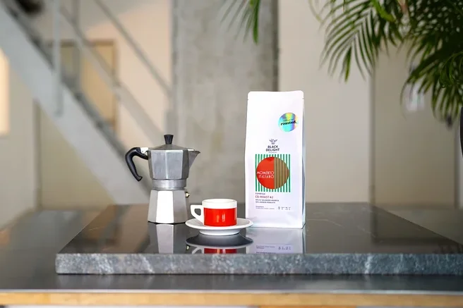 Auf einem Tisch stehen eine Kaffeemaschine, eine Tasse und eine Packung Black Delight x roasted. - Momento Italiano Co-Roast #2 Espresso.