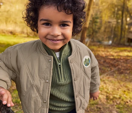 Ein lächelnder kleiner Junge mit lockigem Haar steht in einem sonnigen Wald neben einem Baum und trägt eine beige Steppjacke über einem grünen Rippstrickpullover.