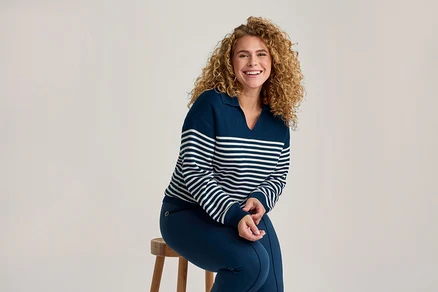 Lachende Frau mit Locken, die einen marineblauen und weiß gestreiften Pullover und marineblaue Hosen trägt, sitzt auf einem Holzschemel.
