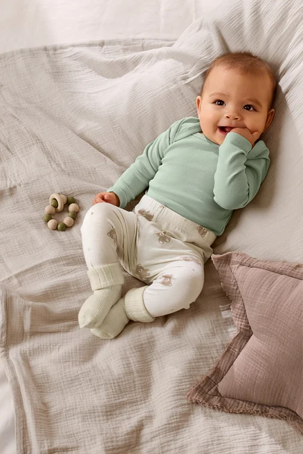 Lächelndes Baby in hellgrünem Body und weißer Hose mit Bärenmuster, das auf einer hellen Decke liegt. Daneben liegt ein Holzbeißring.