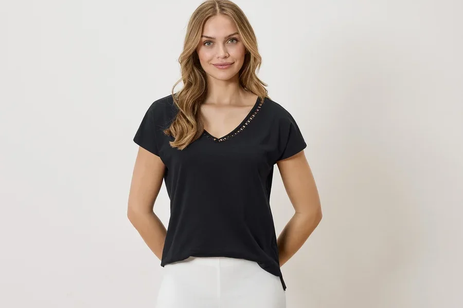Frau mit blonden Haaren trägt ein schwarzes T-Shirt mit V-Ausschnitt und weißen Hosen vor einem neutralen Hintergrund.